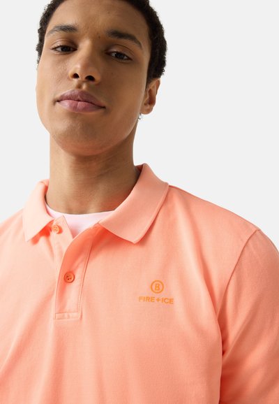 Mladý muž ve světle meruňkovém polotriku s límečkem a dvěma knoflíky, s nápisem "FIRE + ICE" na hrudi, na jednobarevném světlem pozadí.