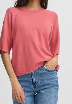 Kobieta ubrana w różowy sweter z krótkim rękawem i niebieskie jeansy, jedną ręką w kieszeni, drugą opartą po boku.