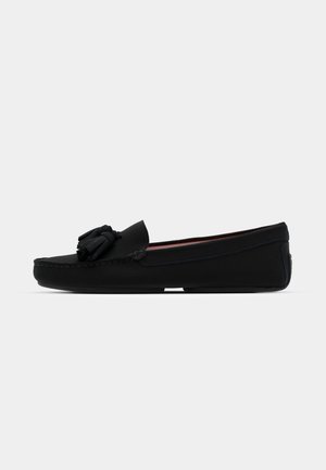 Schwarzer Mokassin-Shoe aus glattem Material, mit einem dekorativen Quasten, genähten Details und flacher Sohle.