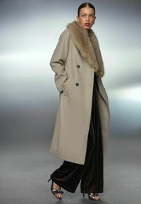 Dubbelzijdige beige jas met een nepbontkraag, voorzien van zijzakken en een lange, vloeiende silhouet, gecombineerd met donkere fluwelen broek en hoge hakken.