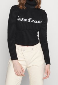 Czarny prążkowany krótki golf z napisem „LetsTrans” w białym kolorze. Do tego jasne spodnie z wysokim stanem.