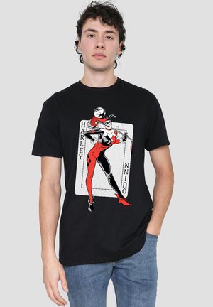 Young man wearing a black t-shirt with a red and black Harley Quinn graphic and blue jeans, standing against a plain gray background.