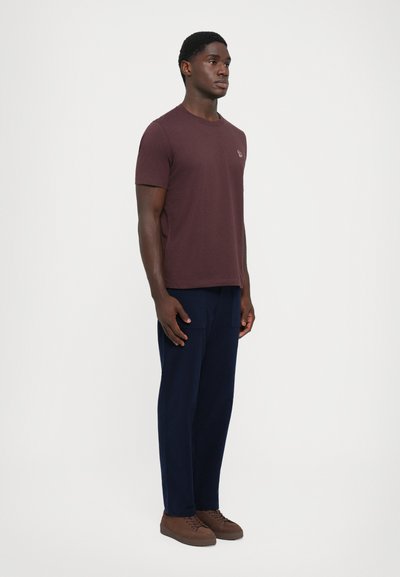 Man die zijwaarts staat, gekleed in een maroon t-shirt, marineblauwe broek met voorzakken en bruine veterschoenen op een witte achtergrond.