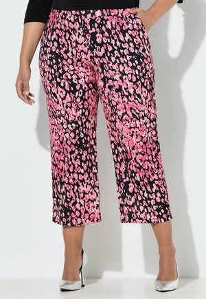 Kvinde iført sorte og pink bukser med abstrakt leopardprint og sølvfarvede høje hæle, stående op ad en hvid væg.