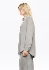 Graues Denim-Hemd mit langen Ärmeln, lockerer Passform und abgerundetem Saum. Mit Knopfdetails und subtiler Textur.