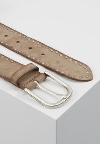 Suede opasek ve světle hnědé barvě se stříbrnou kovovou sponou, zdobený rovnoměrně rozloženými stříbrnými hroty podél pásu, s několika dírkami pro nastavení.