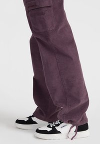 Purple corduroy pants with a relaxed fit, featuring front pockets and a soft texture, paired with black and white sneakers.