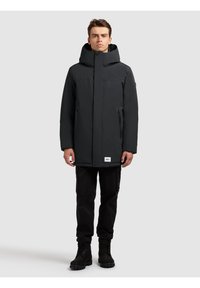 Czarna wodoodporna kurtka z wysokim kołnierzem i kapturem, wyposażona w dwie przednie kieszenie na zamek oraz metkę z logo przy dolnym brzegu. Do kompletu czarne spodnie cargo.