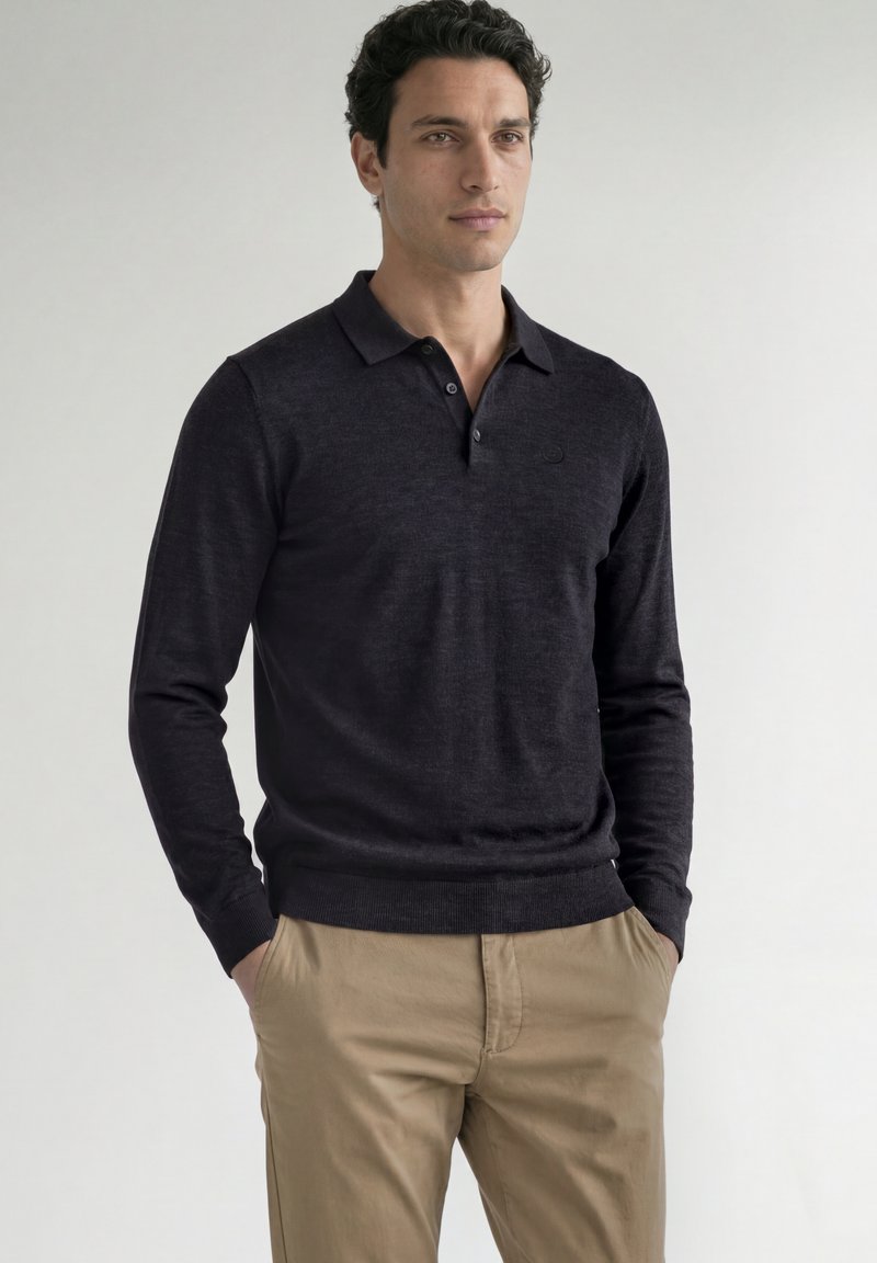 Homme aux cheveux bouclés foncés portant un polo noir à manches longues et un pantalon beige, debout les mains dans les poches devant un fond uni.