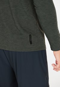 Tummanvihreä pitkähihaisessa paidassa on teksturoitu kangas ja näkyvä musta harjoitusmerkki sivussa. Yhdistettynä tummansinihin shortseihin.