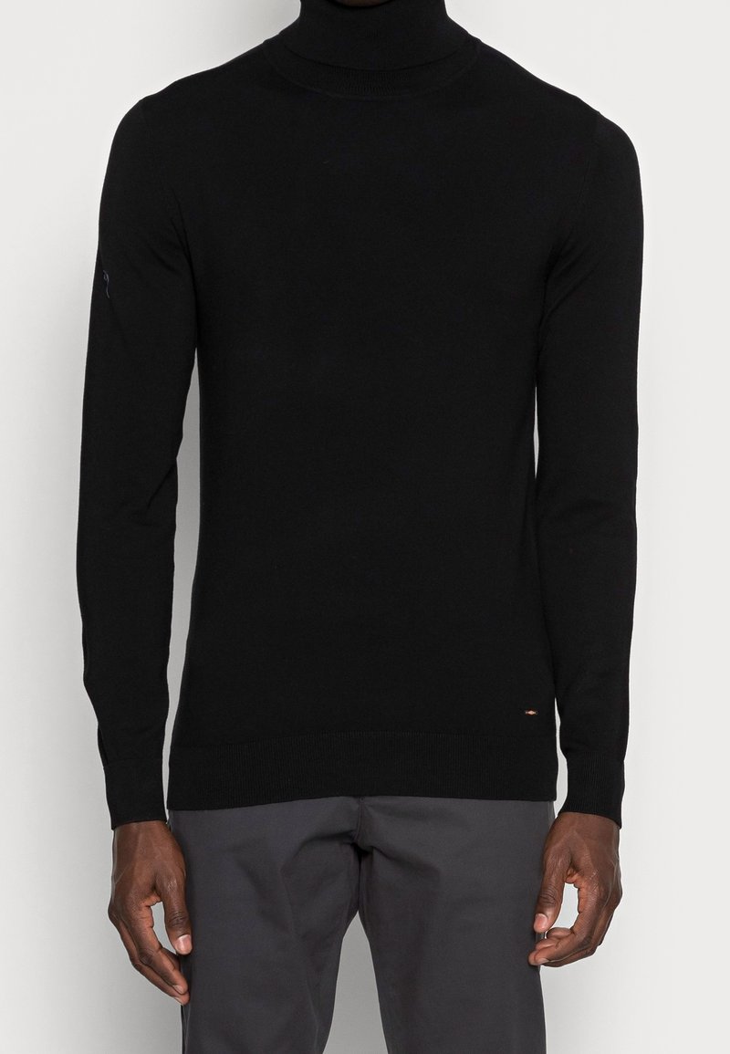 Svart polotröja gjord av fint stickat material, med långa ärmar, ribbade muddarochn en diskret logodetalj vid nederkanten.