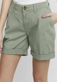 Shorts en coton verts avec une fermeture à bouton, des poignets roulés et des poches latérales. Le tissu a une texture lisse et un ajustement décontracté.