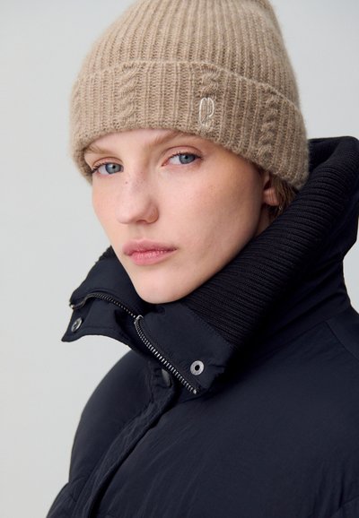 Giacca a vento nera con colletto alto, dotata di zip argentata e bottoni a pressione. Berretto a maglia beige con texture a coste e logo discreto.