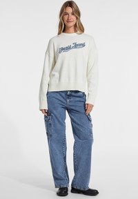 Pulover crem supradimensionat cu logo-ul "Guess Jeans", cu manșete și tiv cu nervuri; asortat cu pantaloni cargo din denim albastru, cu croi lejer și cizme negre.