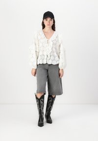 White, eyelet blouse with ruffled sleeves, paired with gray denim shorts. Black knee-high boots with silver embroidery. Black cap.