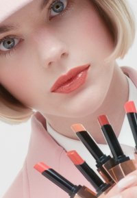 Roze lipsticks in verschillende tinten tentoon gesteld in een waaierformatie. Het model heeft een lichte huid en een zachte roze outfit, wat een frisse make-up look vastlegt.