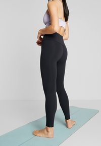 Leggings neri a vita alta realizzati in tessuto elastico, con una texture liscia e un design aderente. Indossati con un reggiseno sportivo lilla.