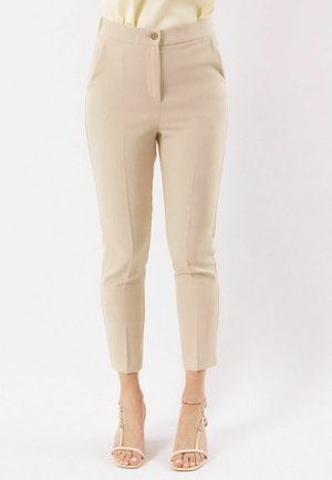 Pantaloni beige a sigaretta alla caviglia indossati con sandali beige con tacco alto, ritagliati dal metà busto ai piedi su uno sfondo neutro.