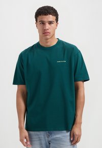 Teal katoenen t-shirt met een ronde hals en korte mouwen. Bevat een klein "HANG ELEVEN" logo in het geel op de linkerborst. Eenvoudig ontwerp.