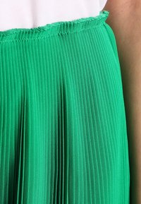 Groene geplooide rok met een golvende zoom, gemaakt van lichte stof. Close-up toont verticale plooien en een soepele textuur bij de taille.