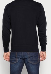 Svart sweatshirt med långa ärmar, ribbade muddar och fåll, med en slät textur och subtila sömnadsdetaljer. Bärs med mörkblå jeans.