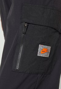 Los pantalones deportivos negros presentan una tela texturizada con un bolsillo con cremallera en el muslo. En el bolsillo se puede ver un colorido parche del logo de Nike.