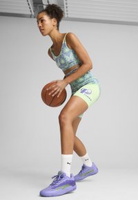 Sportlerin in einem gemusterten blauen Sport-BH und limonengrünen Shorts, die einen Basketball hält. Sie trägt lila Turnschuhe und weiße Socken.
