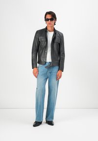 Chaqueta de cuero negra con múltiples bolsillos frontales, llevada sobre una camiseta blanca. Jeans de pierna ancha de color azul claro y zapatos negros completan el atuendo.