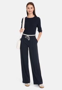 Marineblauer gerippter Pullover kombiniert mit hochgeschnittenen, locker sitzenden Hosen. Weiße Kordelzugbund und cremefarbige Umhängetasche mit dekorativem Riemen.