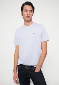 Mlad moški v svetlo modri majici Calvin Klein in temnih kavbojkah, stoji z eno roko v žepu ob enobarvnem belem ozadju.