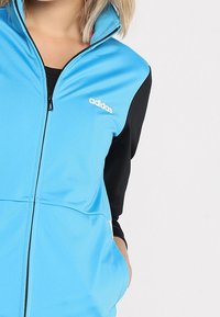 Femme portant une veste Adidas bleue zippée par-dessus un haut noir à manches longues, la main dans la poche de la veste, sur fond uni.