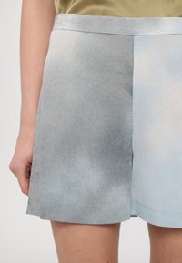 Gros plan d'une personne vêtue d'un short ample dégradé bleu clair et d'un haut beige, montrant la main gauche et les jambes supérieures sur un fond uni.