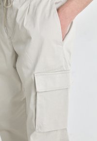 Beige Cargo-Hosen aus weichem, leichtem Stoff mit seitlichen Taschen, Kordelzugbund und genähten Details auf den Taschenklappen.