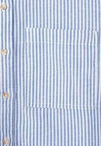 Poche de chemise à rayures verticales bleues et blanches avec des boutons beige le long de la patte de boutonnage sur le côté gauche.