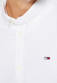 Valkoinen napitettava paita, jossa on kaulus ja muoviset napit. Huomattava logo punaisena, valkoisena ja sinisenä vasemmassa rinnassa.