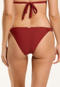 Mélyvörös anyagú bikini alsó, sima textúrával, alacsony szabással, összegyűrt oldalsó megkötőkkel és egy kis arany kiegészítővel.