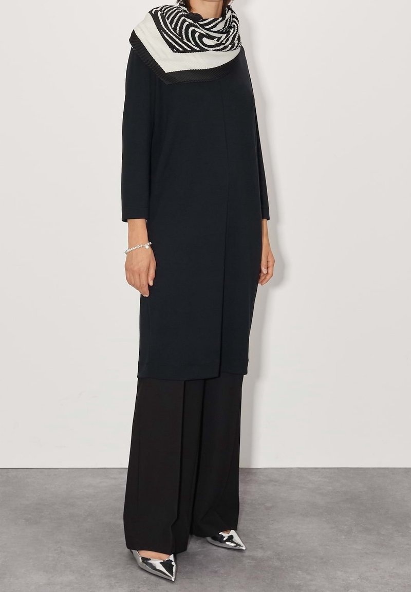Femme portant un long manteau noir, un pantalon noir large, des chaussures argentées pointues, une écharpe à motif noir et blanc, et un bracelet argenté.
