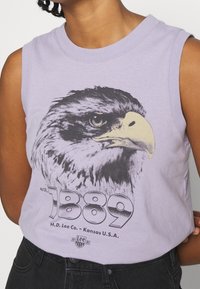 Vaaleanvioletti hihaton t-paita, jossa on yksityiskohtainen mustavalkoinen kotka-kuvake, teksti "est. 1889" ja brändin nimi.