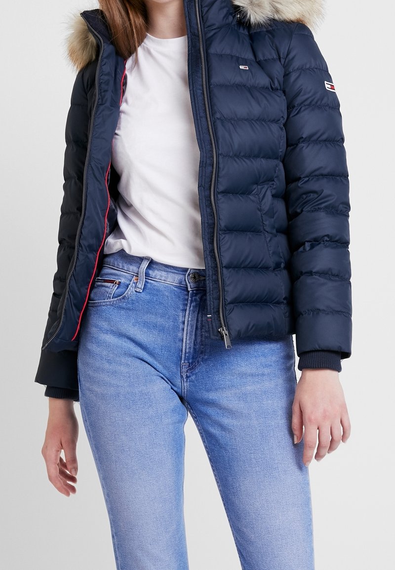 Námořnická péřová bunda s odnímatelnou kapucí s kožešinou, zapínáním na zip a červenými detaily na podšívce. Nošena přes bílou košili a světlé modré džíny.