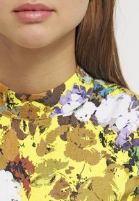 Top con motivo floreale su base gialla; presenta un collo alto e un tessuto testurizzato con tonalità di fiori viola, marroni e bianchi.