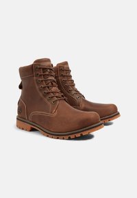 Botas de tobillo de cuero marrón con costuras beige contrastantes, ojales de metal y suelas de goma resistentes. Presenta detalle del logo en el lateral.