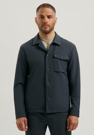 Man met kort haar en baard, gekleed in een donkerblauwe, met knopen sluitende jas met een borstzak en een beige shirt eronder, staande tegen een eenvoudige achtergrond.
