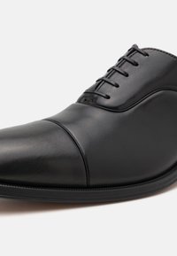 Svart lær dressko med avrundet tå, glatt tekstur, buet design og seks snøringshull. Har en subtil sømdetalj ved tåen.