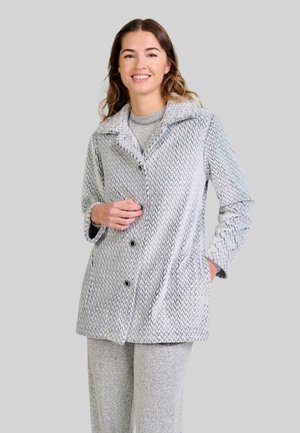 Mujer sonriente que lleva una chaqueta abotonada gris con estampado sobre unos pantalones gris a juego, de pie frente a un fondo gris liso.