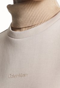 Suéter de color beige con cuello alto, hecho de una tela suave, que presenta un cuello acanalado y un sutil logo de Calvin Klein grabado en el dobladillo.