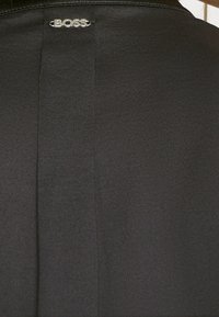 Tissu noir avec une étiquette logo "BOSS" argentée cousue près du centre en haut, montrant la texture et les détails de la couture verticale.