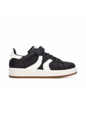 Zapatilla negra de corte bajo con rayas blancas en zigzag, cierre de velcro, cordones negros, detalle blanco en el talón y suela marrón.
