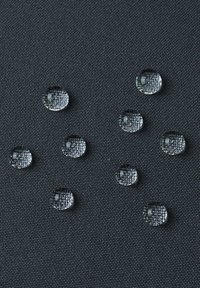 Gocce d'acqua si distribuiscono su una superficie di tessuto scuro e strutturato, mostrando una varietà di dimensioni e proprietà riflettenti.