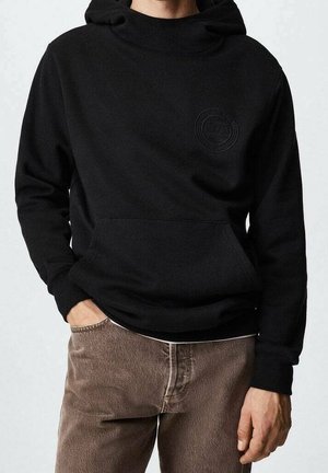Personne portant un sweat à capuche noir avec une poche avant et un logo discret sur la poitrine, associée à un pantalon marron, main dans la poche.