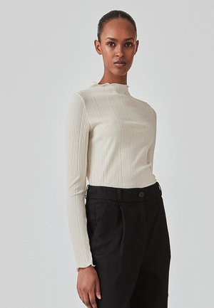 Top a maniche lunghe in maglia a coste color crema con un collo alto e un leggero volant. Abbinato a pantaloni neri su misura a vita alta.